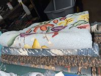Three rugs folded and stacked, showing braided patterns on two rugs and the edge of the floral hand hooked rug on top.