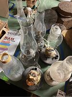 Collection of oil lamp bases and various glass chimneys shown on table, includes one electric lamp and several black metal bases with old wicks.