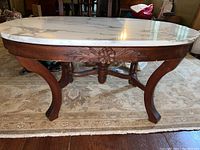 Full view of the wood table with an oval white marble top and carved wooden apron with floral motifs, standing on a rug over wood floor.