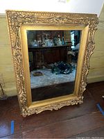 Full view of the rectangular, gold-tone ornate framed mirror showing its size and condition in a room with wood walls and furniture reflected in the glass.