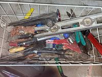Top-down view of a wire basket containing various hand tools such as pliers, scrapers, knives, files, and a metal spirit level with two bubble vials.