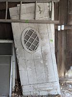 Two wooden doors leaning against a barn wall. One door has an oval lattice-style window, both have worn white paint.