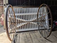 Front view of two rusty metal wagon wheels connected by an axle shaft placed on wooden floor with a white fence background.