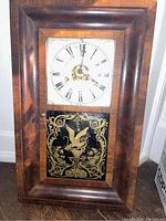 Front view of the vintage wall clock showing the wood frame, white dial with Roman numerals, and decorative gold and black glass panel with eagle motif.
