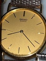 Close-up of Seiko Quartz gold-tone watch face with black indices and leather band, showing wear and scratches.