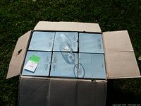 Open cardboard box containing 6 sealed boxes of 4 wine glasses each, total 24 glasses. One glass is placed on top inside the box showing clear glass, bowl shape, stem, and base.
