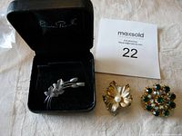 Three brooches displayed including a sterling silver floral spray brooch in a black velvet box, a leaf brooch with faux pearl, and a colorful rhinestone brooch.
