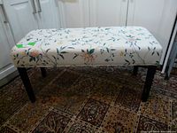 Side view of bench showing floral upholstery with beige background and wooden legs on a patterned carpet.