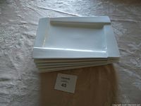Stack of six white rectangular dinner plates on a light patterned tablecloth, showing side view and thickness of plates.