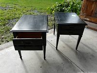 Pair of black wood end tables with drawers. Tops show scratches, one drawer partially open with handle detached inside.