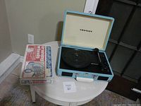 Blue Crosley record player in suitcase form with tonearm and turntable visible beside white box with 'training wheels' label.