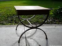 Side table with wooden top and metal legs standing on concrete surface outdoors with grass and leaves in background, shown from front angle.