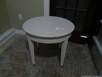 Front and top view of small round white painted wood side table showing the full table with round top and four legs on a carpeted floor.