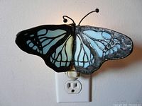 Butterfly stained glass nightlight plugged into electrical outlet, showcasing left side wing with translucent light blue glass and black metal outlines.