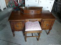 Front view of vintage dressing table showing center top drawer and two side drawers with decorative handles. Upholstered floral bench is in front.