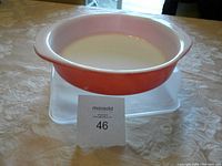 Pink and white vintage Pyrex baking dish placed on a beige tablecloth, elevated on a clear plastic container (not included). Front view showing the full dish profile including handles.