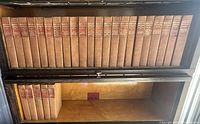 27 uniform brown cloth volumes with red gilt spine labels arranged on two shelves of a wooden bookcase