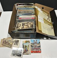 Box filled with numerous vintage postcards standing upright inside a black storage box, some postcards laid out in front showing various designs and text, including a black and white postcard with an indoor museum scene, and a Roma postcard.