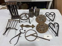 Overview of 13 antique metal kitchen and household items including trivets, containers, tongs, and other metal tools.