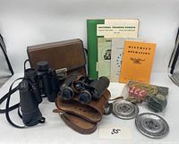 Two pairs of binoculars with cases, Boy Scout manual booklets and papers, pewter medallions, lanyard, and patch laid out on a white surface.