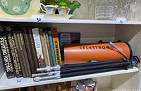 Shelf displaying orange Celestron telescope tube with black legs of a tripod below it, alongside several hardcover and paperback astronomy books.