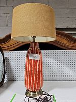 Full view of the orange ceramic mid-century style lamp showing vertical white stripes, brass base, beige fabric shade, power cord coiled on the table.