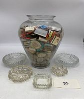 Front view of large glass jar filled with colorful matchbooks with five clear glass ashtrays arranged around it on the table.
