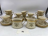 Full view showing 12 tea cups stacked on matching saucers along with cream pitcher and sugar bowl, all with gold floral decoration.