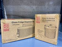 Photo of boxed glass fridge dispenser 5L and boxed glass ice bucket with bamboo lid positioned side by side on shelving unit, showing labels and product illustrations on boxes.