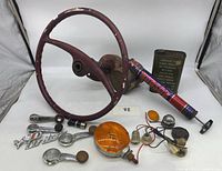 Photo shows red metal tractor steering wheel with rusted base, multiple small car parts including Pinto badges, amber and clear vehicle lights, a green Hudson Pest Control can, and a red cylindrical item.