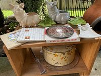 Wide shot of two ceramic pig planters, trivets, cake pan top, enamel pot, and other items arranged on wooden shelf outdoors.