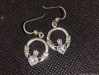 Close-up photo of pair of sterling silver Claddagh earrings with clear heart-shaped topaz stones on black textured surface.