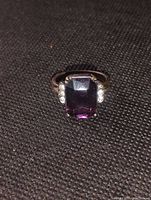 Close up photo of the silver plate faux amethyst Art Deco style ring lying flat on dark textured fabric, showing the rectangular purple faux amethyst center stone and side clear artificial diamond clusters.