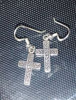 Pair of sterling silver Celtic cross earrings lying on a textured black surface, showing front and side view of hooks and crosses with intricate knotwork design.