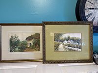 Two framed English cottage landscape prints displayed side by side. One is smaller in gold frame, the other larger in bronze frame.