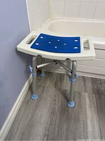 Bath seat positioned near bathtub, shows top view of blue slip-resistant seat and side handles for support, plastic frame with metal legs and rubber feet.