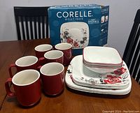 Photo showing six solid red mugs, stack of square soup/cereal bowls, salad plates, and dinner plates with floral red/black pattern, plus unopened boxed 16-piece Corelle Boutique set.