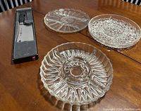 Three different clear cut glass divided serving plates beside a boxed Henckel cutting utensil, displayed on wooden table