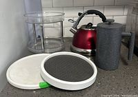 Red metallic whistle kettle, granite-look coffee press, two-tier clear acrylic turntable, and two single clear turntables shown on kitchen counter.