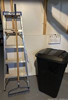Wide view of ladder, garbage pail, and garden rake arranged in basement with white wall background.
