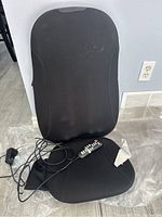 Front view of black portable chair massager with wired remote and power cord laid on floor, showing wear marks on fabric and contoured padded design