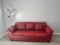 Red leather couch with visible wear, three seat and back cushions, rounded armrests, positioned near wall corner.