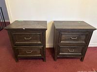 Pair of dark wooden side tables shown from front, each with two drawers and metal handles, placed on red carpet against white wall.