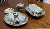 Photo showing three silver-plated serving dishes with lids, a silver-plated sugar bowl on copper, and a small round silver-plated Rogers dish on a wooden table.