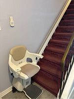 Wide angle showing entire stair chair lift installed on the staircase with red carpet and beige cushioned seat.
