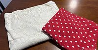 Two folded tablecloths on a wooden table; red with white polka dots and white lace