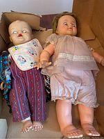 Two antique dolls in cardboard box, one wearing striped outfit and bib, the other in pink dress.