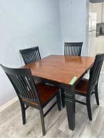 Photo showing square wooden table with medium brown top and black painted legs, surrounded by four black slatted-back wooden chairs with brown seats.