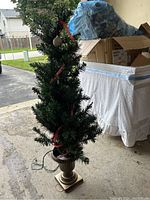 Single artificial Christmas tree decorated with red ribbons and Christmas balls, mounted on ornate urn-style planter stand.