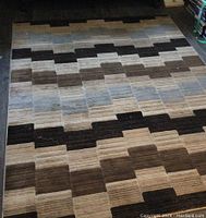 Full view of the rectangular area rug showing overall geometric stepped block pattern in neutral tones of beige, brown, gray, and tan.